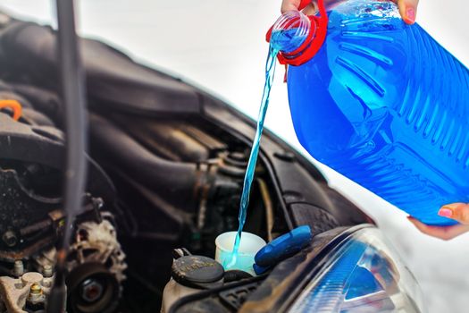 Woman pouring antifreeze car screen wash liquid into dirty car from blue anti freeze water container.