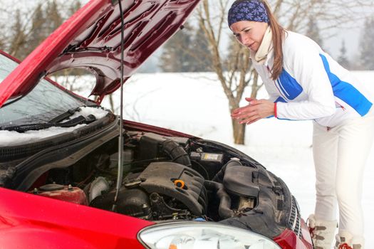 Lonely worried young woman in sport jacket looking on engine in opened car hood trying to fix her broken red car on winter snowy day.