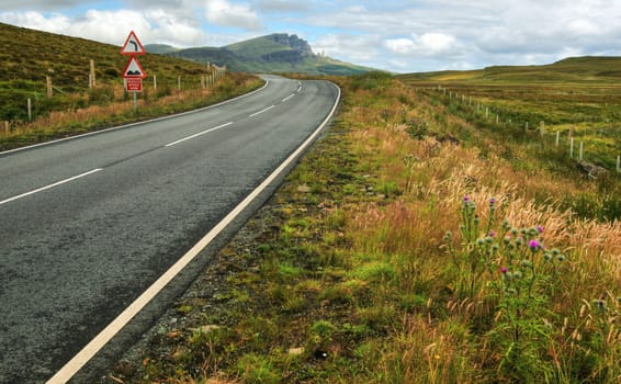 Wide shot of empty road, curve, bumps and reduce speed now sign with Old Man of Storr mountain in the background. Isle of Skye, Scotland, United Kingdom.