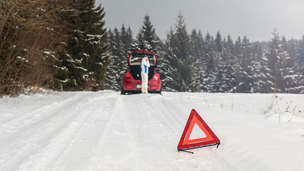 Bright red triangle on a snowy road warning other drivers with woman standing next to broken car in forest, calling repair service on her mobile phone.