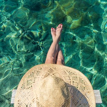 Graphic image of top down view of woman wearing big summer sun hat relaxing on small wooden pier by clear turquoise sea.