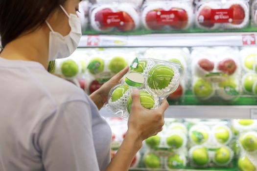 Women long hair wear protective face mask hands holding green apples ipackage. Asian female shopping at supermarket, Grocery to buy some food. New normal after covid-19 concept.