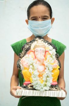 Girl Kid with medical mask holding Ganesha Idol on isolated background - concept of vinayaka Chaturthi festival celebrations during coronavirus or covid-19 pandemic.