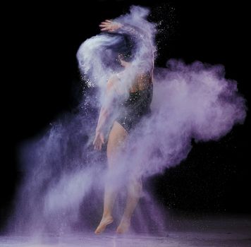 beautiful caucasian woman in a black bodysuit with a sports figure is dancing in a purple cloud of flour on a black background