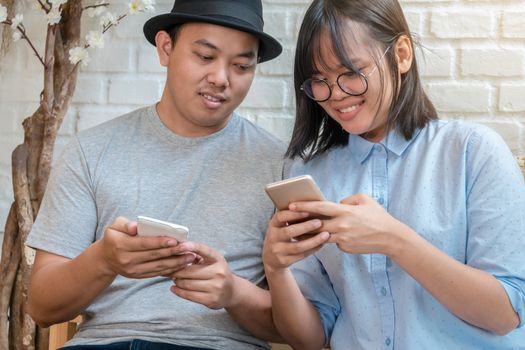 Asian Young Couple talking and using the smart mobile phone with happiness action in modern coffee shop or workplace or co-working space or modern office, lifestyle and leisure concept