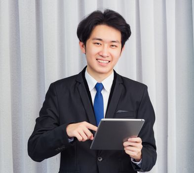 Work from home, Asian young businessman wear suit video conference call or facetime he smiling using smart digital tablet computer and point finger on the screen at home office, looking to camera