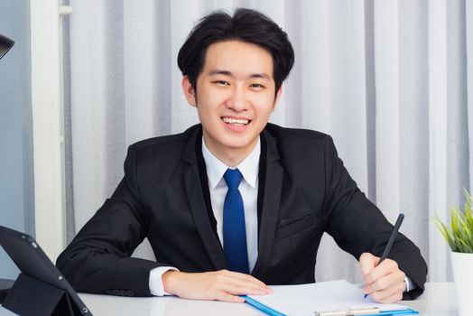 Work from home, Asian young businessman video conference call or facetime he smiling looking to camera sitting on desk using smart digital tablet computer and write notebook information at home office