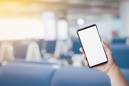 Hand holding black smartphone or mobile phone with white blank screen on blurry blue seats at airport background. Travel concept.