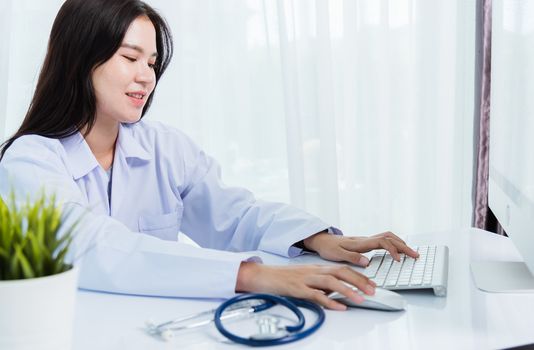 Asian beautiful female doctor smiling wearing doctor's uniform with stethoscope typing on keyboard desktop computer looking to monitor on the desk at hospital office, Medical healthcare concept