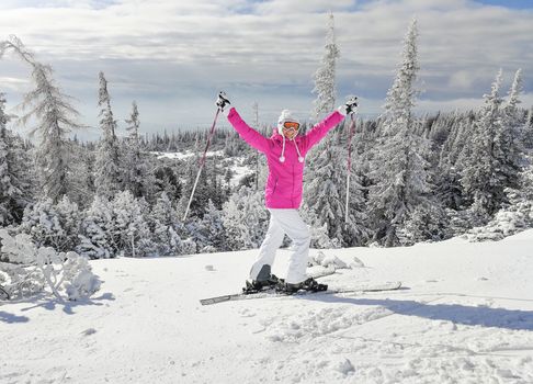 Young woman in pink ski jacket with skis on her feet holding ski poles up, in happy pose smiling during nice sunny winter day. Strbske pleso ski resort, Slovakia.
