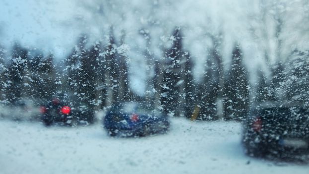 View from inside car to snow flakes on windshield with blurred cars in parking lot in background. Winter weather driving conditions.