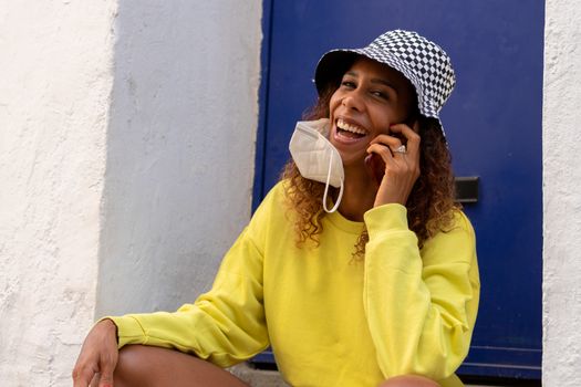 Portrait of an attractive young black woman sitting and talking with the mobile phone outdoors with her mask off. Concept communication