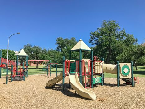 Colorful urban playground at public park surrounded by large trees in downtown Dallas, Texas, America. Empty recreation place in hot summer day with sunny clear blue sky.