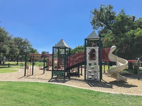 Colorful urban playground at public park surrounded by large trees in downtown Dallas, Texas, America. Empty recreation place in hot summer day with sunny clear blue sky.