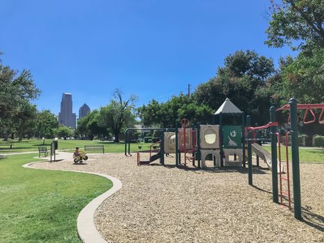 Kid playground with skylines background at public park surrounded by large trees in downtown Dallas, Texas, America. Empty recreation place in hot summer day with sunny clear blue sky.