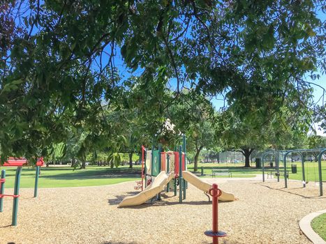 Large tree shade and colorful urban playground at public park in downtown Dallas, Texas, America. Empty recreation place in hot summer day with sunny clear blue sky.