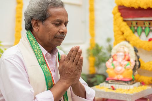 Elder man offering Prayer by namaste gesture in front of Lord Ganesha Idol during Ganesha or vinayaka Chaturthi festvial ceremony at home