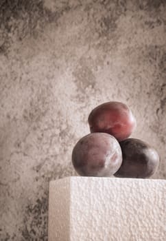 Three large ripe plums on a light podium against a dark grunge background. Front view, copy space