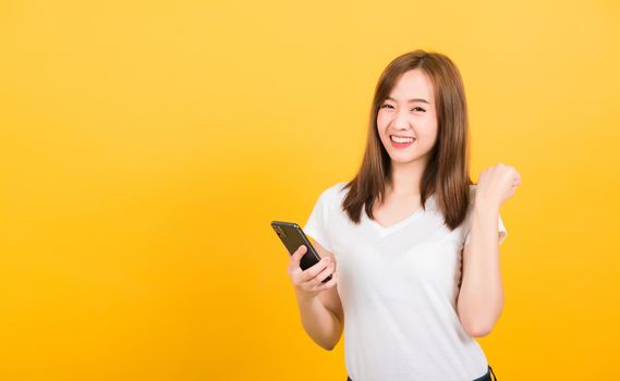 Asian happy portrait beautiful cute young woman teen smiling standing wear t-shirt celebrating win with smart mobile phone looking the phone isolated, studio shot on yellow background with copy space