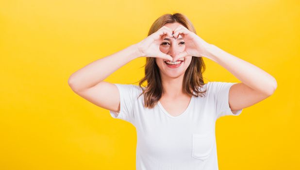 Asian Thai happy portrait beautiful cute young woman smile standing make finger heart figure symbol shape sign with two hands and looking camera, studio shot isolated yellow background with copy space