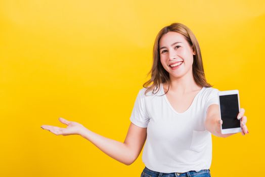 Portrait Asian Thai beautiful happy young lifestyle woman stands holding mobile phone and show something on side hand looking to camera, studio shot isolated on yellow background, with copy space