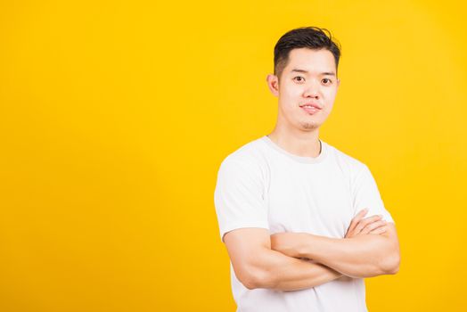 Portrait happy Asian handsome young man smiling standing wearing white t-shirt folded or crossed arm he looking to camera, studio shot isolated yellow background