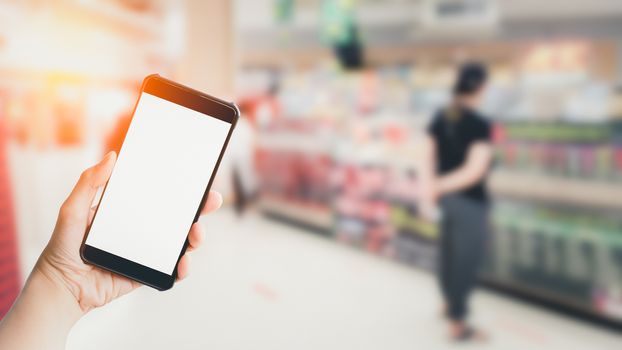 Shopping online concept. Hand holding black smartphone and white blank screen in blurry at shopping or department store background. Mock up shopping.