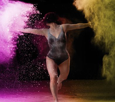 beautiful caucasian woman in a black bodysuit with a sports figure is dancing in a pink yellow cloud of flour on a black background