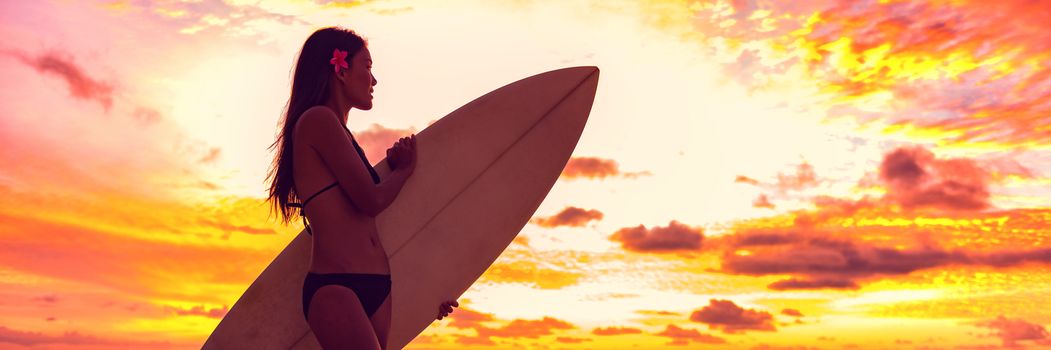 Surfer bikini girl on Hawaii beach holding surf board watching ocean waves at sunset. Silhouette of Asian sport woman over landscape, sky and clouds background. Summer vacation lifestyle.