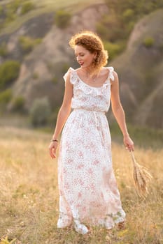 Beautiful tender girl in a white sundress walks at sunset in a field with a spikelet bouquet