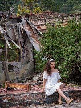 Beautiful young woman in light dress sitting in front of the ruins of a ghost town