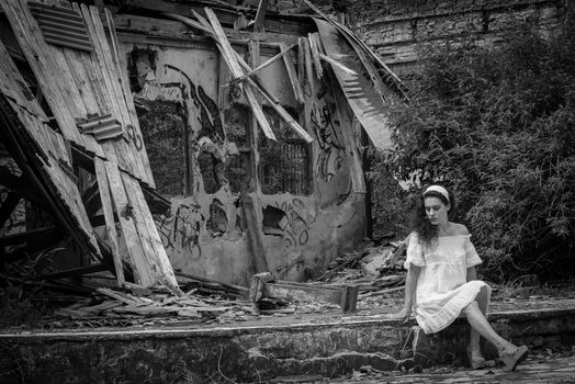 Beautiful young woman in light dress sitting in front of the ruins of a ghost town