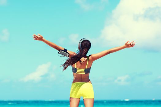 Fitness healthy lifestyle runner exercise success. Happy woman running with open arms in freedom listening to music with earphones and smartphone armband at beach ocean with sunshine filter.
