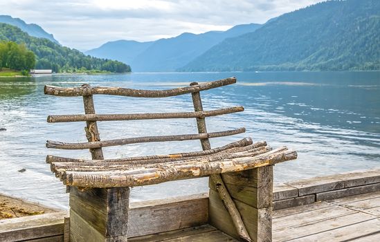 view of a wooden bench made of tree branches, which stands on a boat pier against the backdrop of a picturesque mountain landscape with a river, lake