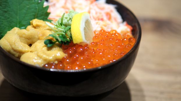 Close-up images of japanese seafood rice bowl or kaisendon sashimi donburi which have fresh ogura, uni, giant crab and topping with lemon on rice in the black bowl on the wood table from top view angle camera.