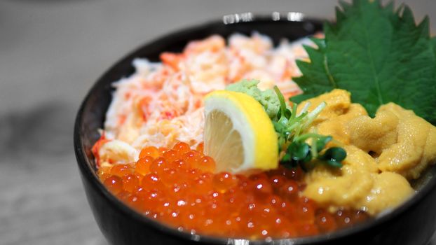 Close-up images of japanese seafood rice bowl or kaisendon sashimi donburi which have fresh ogura, uni, giant crab and topping with lemon on rice in the black bowl on the wood table from top view angle camera.
