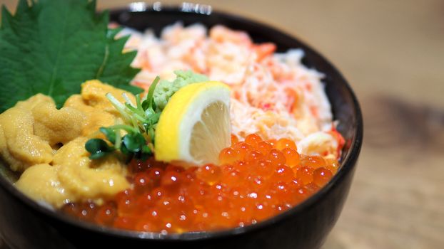 Close-up images of japanese seafood rice bowl or kaisendon sashimi donburi which have fresh ogura, uni, giant crab and topping with lemon on rice in the black bowl on the wood table from top view angle camera.