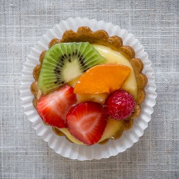 A cake with cream and mixed fruit, resting on linen tablecloth.