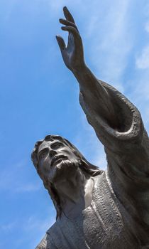 Bronze statue of Jesus looking at the sky with both arms up. On background a blue sky whit white clouds.
