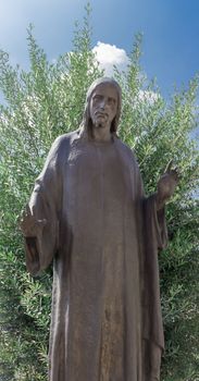 Bronze sculpture of Jesus christ. The Redeemer has his arms open, he wears a tunic. Green plant on the background.