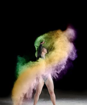 beautiful caucasian woman in a black bodysuit with a sports figure is dancing in a multicolored cloud of flour on a black background
