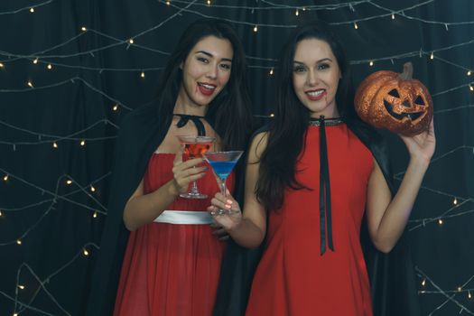 Beautiful woman and friend in Halloween devil costume with carved pumpkin and drinking cocktail. Have fun at a party in nightclub decorated with lights. Halloween celebration