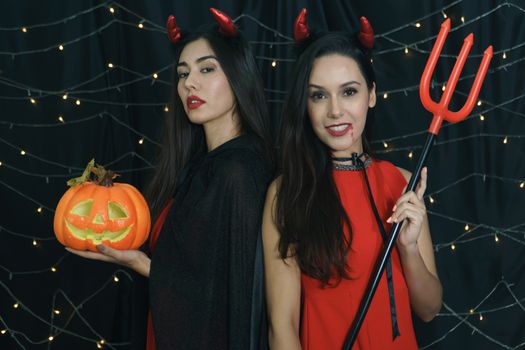 Beautiful woman and friend in Halloween devil costume with carved pumpkin and holy spear. Have fun at a party in a nightclub decorated with lights. Halloween celebration