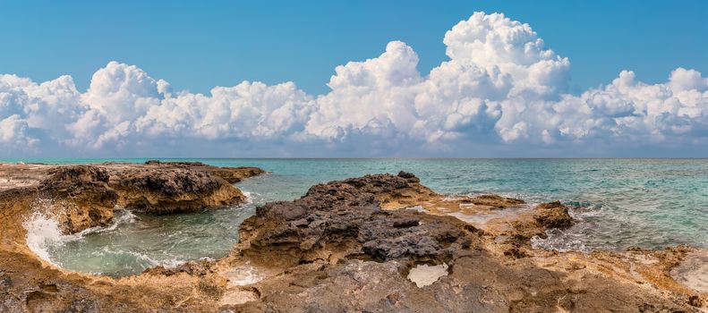 Beautiful panoramic view of a rocky beach in tropics. Turquoise water splashing in the foreground. Scenic blue sky with amazing white clouds as a back