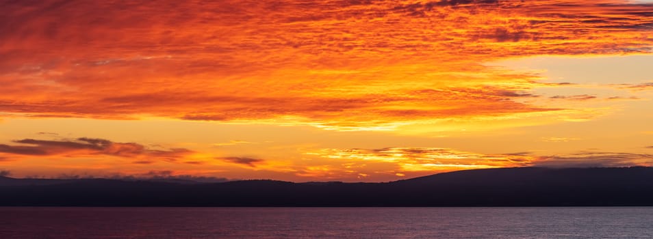 Scenic red-and-orange sunset with a shoreline as a silhouette in the foreground. Amazing panorama.