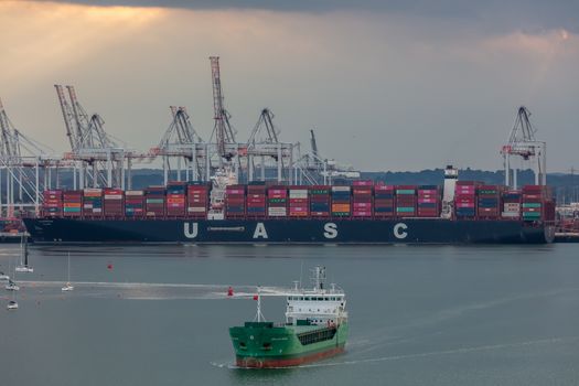 Southampton port, England, UK - June 08, 2020: Aerial view of Southampton port. Sunset. Massive container ship UASC being loaded in the background. Arklow Rogue ship sailing in the foreground.