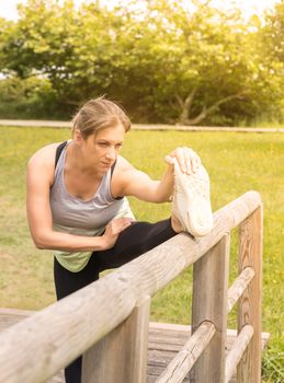 Woman in sportswear stretching her legs on a wooden fence after training in the park.