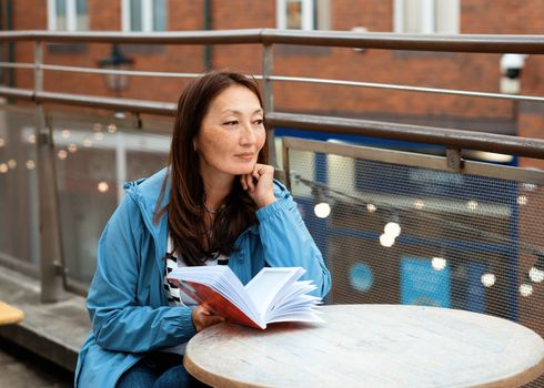 Asian woman in a blue coat sitting in the cafe in the street and reading a book