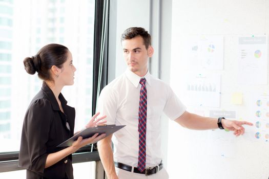 young businessman pointing towards graph and businesswoman holding clipboard with present profit while giving presentation in office, teamwork concept.