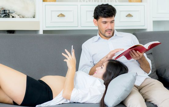 Asian couple family using a tablet at home, the young mother is pregnant lying on sofa looking tablet and husband reading book.